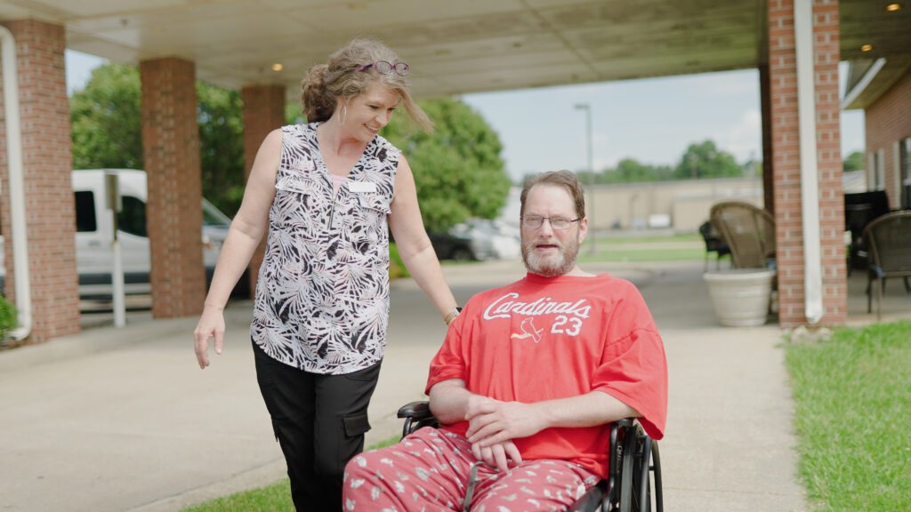A man sits in a wheelchair and smiles at a nurse at a respite care facility